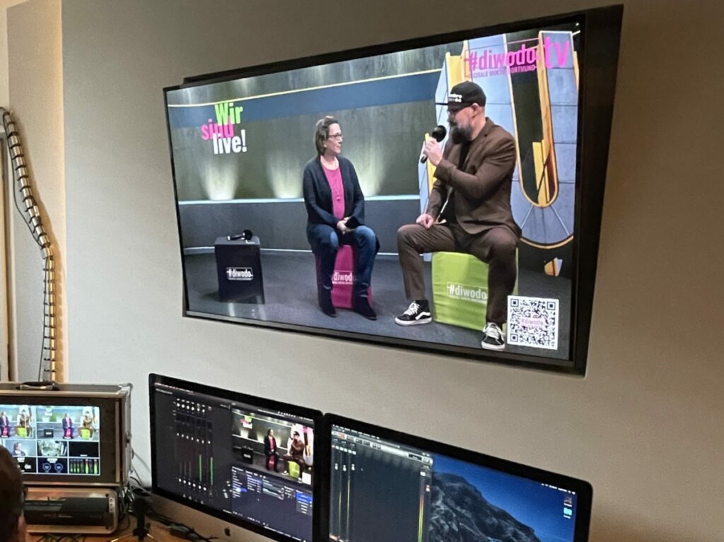 Man sieht ein TV-Studio, auf einem großen Monitor sieht man Maike und Kai im Interview