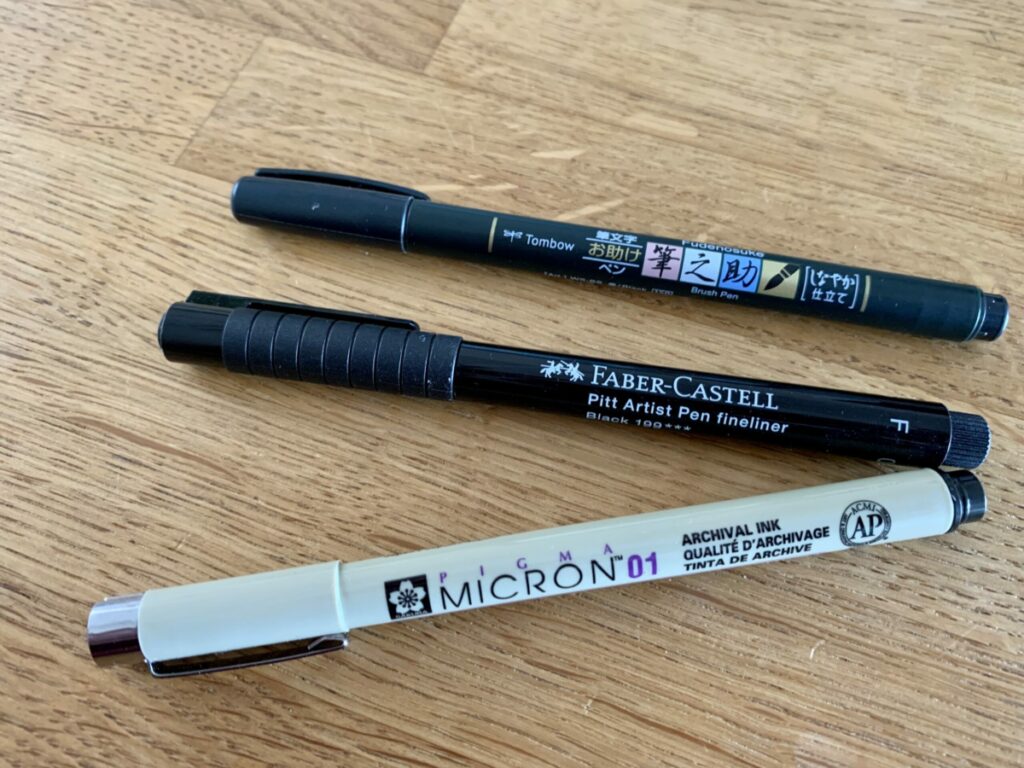 3 Stifte liegen auf einem Holztisch: Tombow Fudenosuke, Faber-Castell Pitt Artist Fineliner und Pigma Micron 01