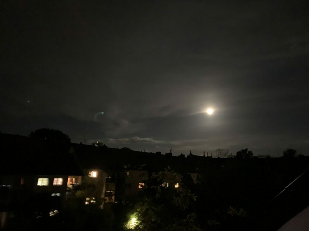 Mond über der Stadt: Obere Bildhälfte: Schwarzer Himmel mit weiß-gelblichem Fast-Vollmond, untere Hälfte: dunkle Häuser mit vereinzelt beleuchteten Fenstern