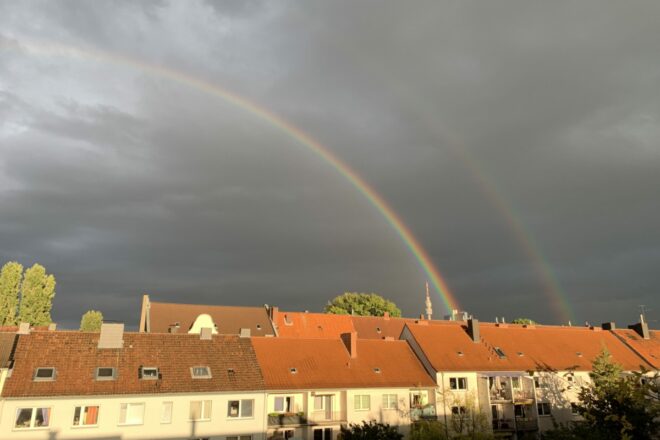 Doppelter Regenbogen: Von der Sonne angestrahlte Häuserreihe vor sehr (!) dunklem Himmel, dahinter zwei Regenbögen