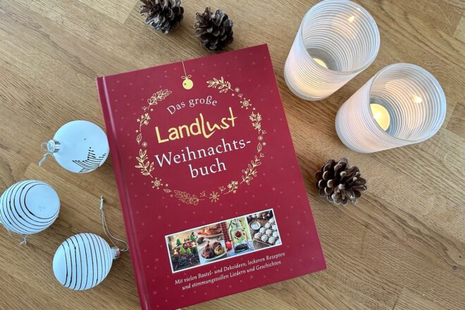 Rotes Buch "Das große Landlust Weihnachtsbuch" liegt auf Holztisch. Darum drapiert zwei weiße Teelichthalter, weiße Christbaumkugeln und Zapfen