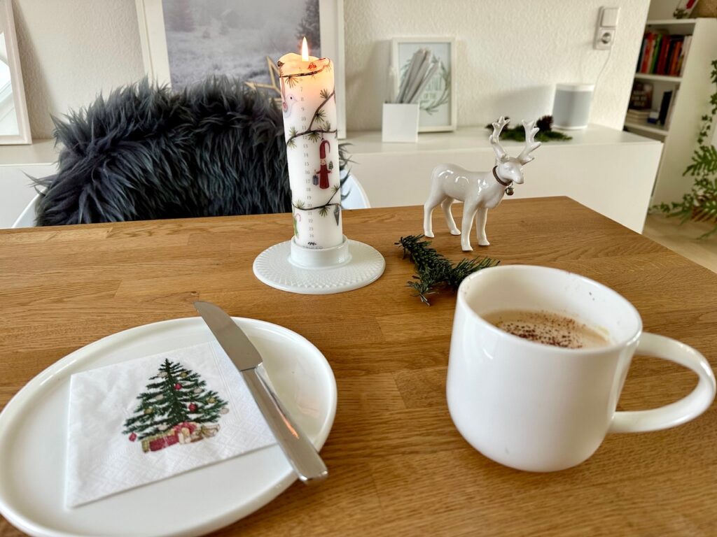 Frühstücksgeschirr (Teller, Tasse und Messer mit Weihnachtsserviette) vor einer Adventskalenderkerze (Kerze mit Weihnachtsmuster und Zahlenreihe von 24 bis 1 drauf), einem kleinen Tannenzweig und einem weißen Porzellan-Rentier.