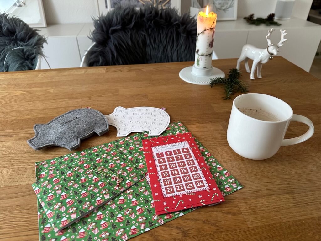 Auf einem Tisch liegen ein großer Umschlag mit weihnachtlichem Muster, eine Karte in dem gleichen Weihnachtsmuuster, ein Umschlag für die Adventskalenderchallenge (Umschlag rot weihnachtlich gemustert mit Felder von 1 bis 24) ein Filzschwein zum Besparen und ein Tracker aus Pappe in der Größe des Filzschweines, auf dem man 30 Fünfen aufgedruckt sind, die abgestrichen werden können.