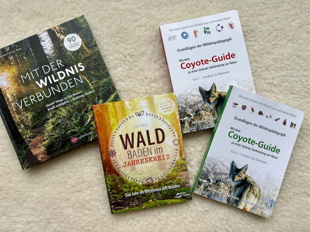 Vier Bücher auf Wollteppich: ""Mit der Wildnis verbunden" von Susanne Fischer-Rizzi, "Waldbaden im Jahreskreis" von Ulli Felber, und "Mit dem Coyote Guide zu einer tieferen Verbindung zur Natur" (Band 1 und 2) von Jon Young