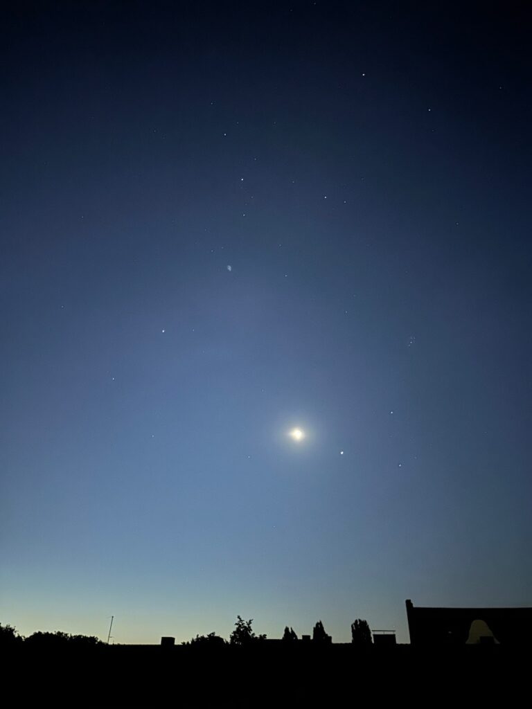 Nachthimmel: Unten die dunkle Silhouette der Dächer, darüber der dunkelblaue Himmel mit zunehmendem Mond und Sternen