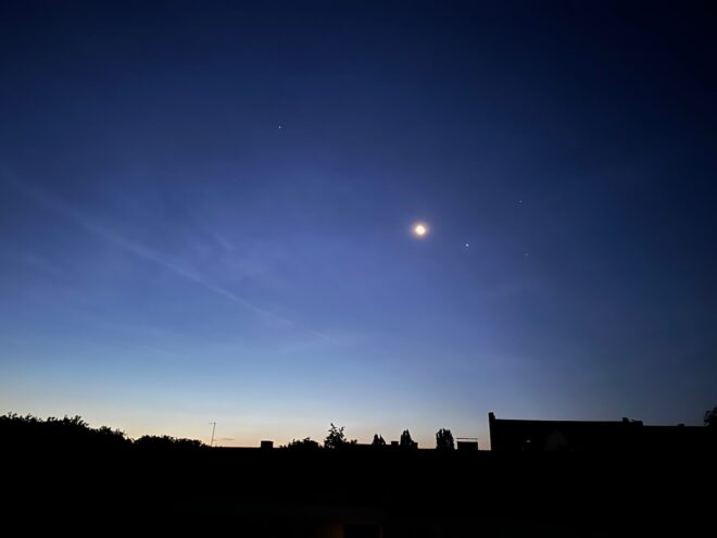 Nachthimmel kurz vor Sonnenaufgang: Unten eine schwarze Silhouette der Häuserdächer, darüber der dunkelblaue Nachthimmel mit einem abnehmenden Mond und diversen Sternen.